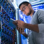 Low angle  portrait of young man using digital tablet standing by server cabinet while working with supercomputer in blue light